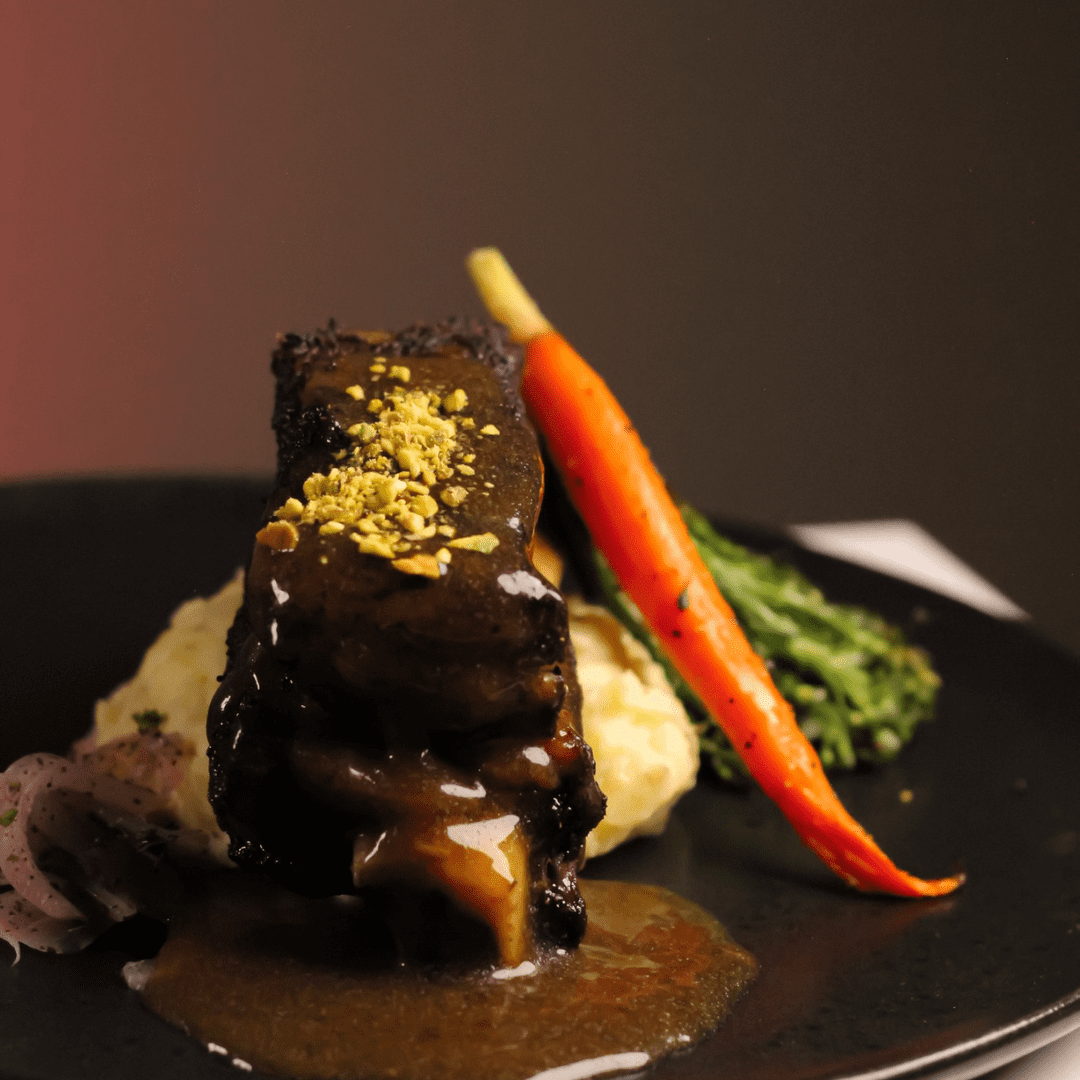 A gourmet dish featuring braised short ribs with a rich sauce, served over mashed potatoes, garnished with crushed pistachios, a whole glazed carrot, and a side of greens on a black plate.