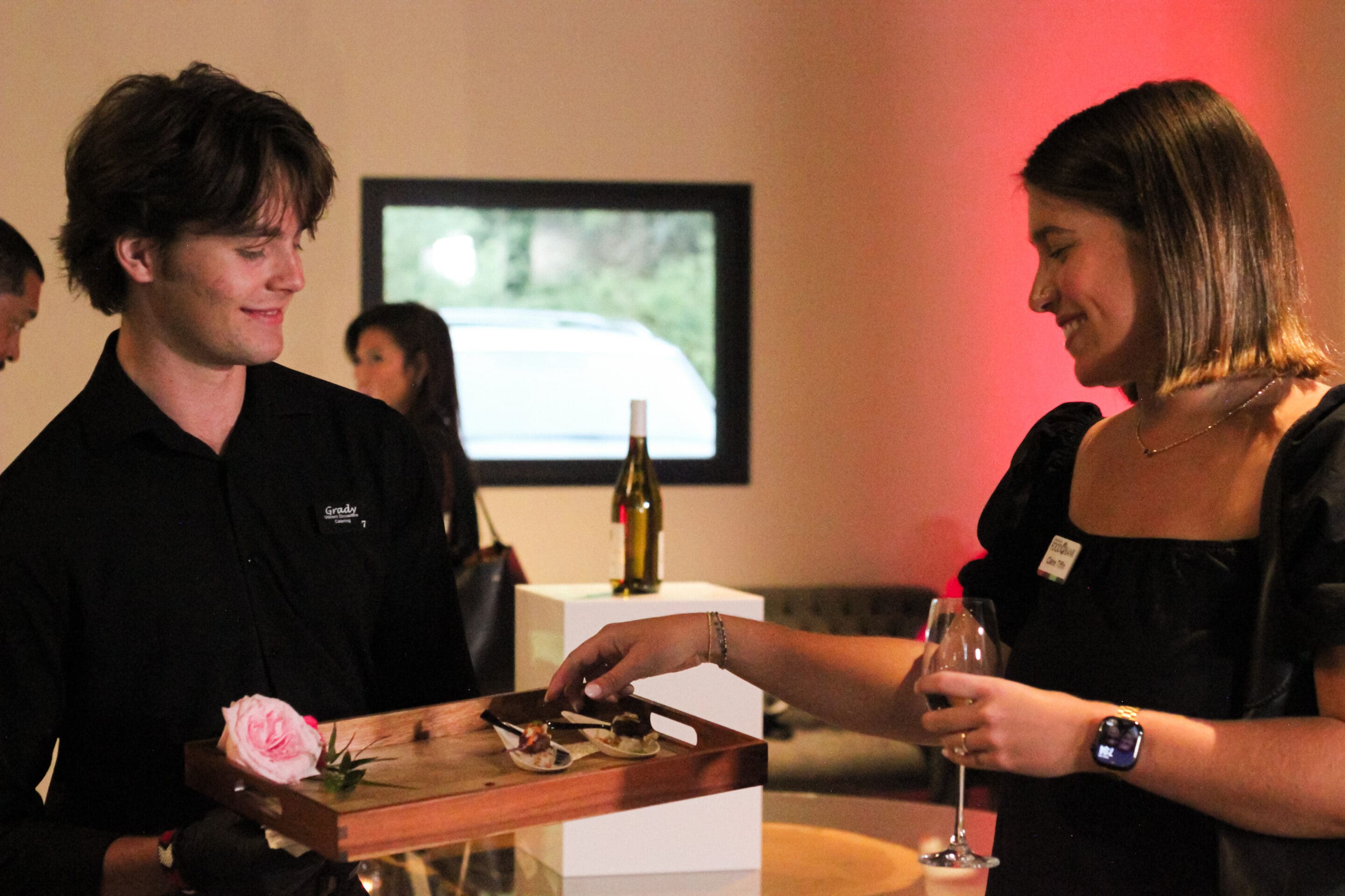 A smiling server holds a wooden tray with appetizers and a pink flower as a woman, holding a glass of wine, reaches for food at a warmly lit indoor event. People and a wine bottle are visible in the background.