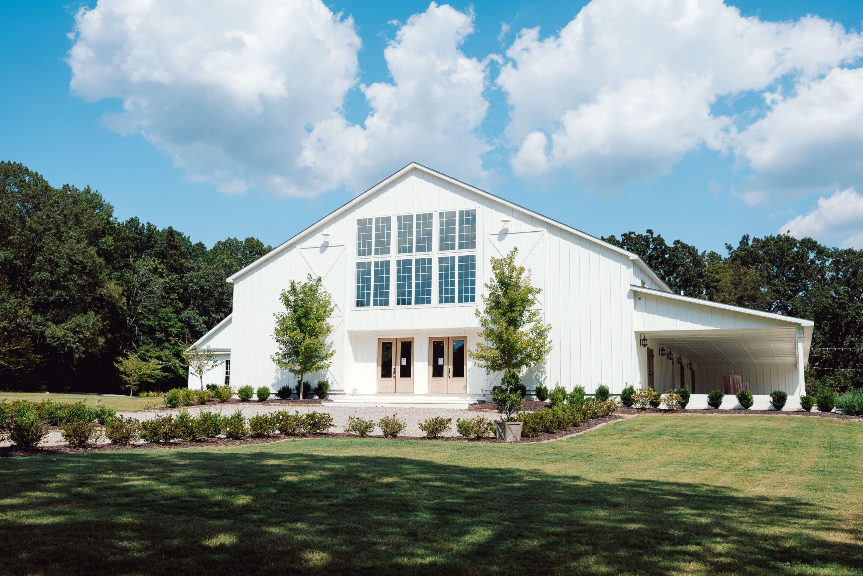 A large white barn-style building with tall windows and double doors sits on a manicured lawn, surrounded by trees under a partly cloudy sky.