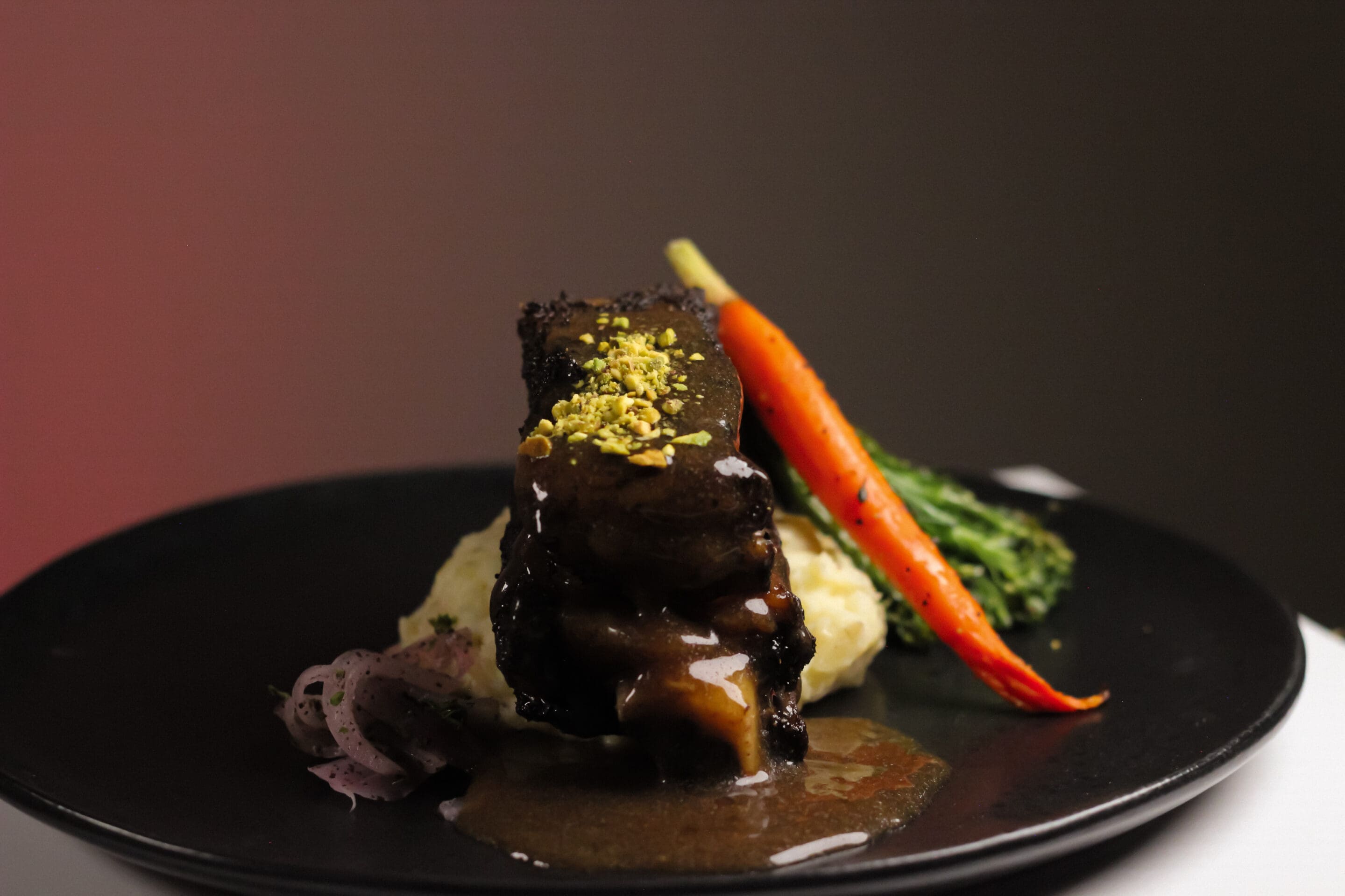 A gourmet dish featuring braised short rib topped with crushed pistachios, served over mashed potatoes with gravy, accompanied by a roasted carrot, broccolini, and pickled onions on a black plate.