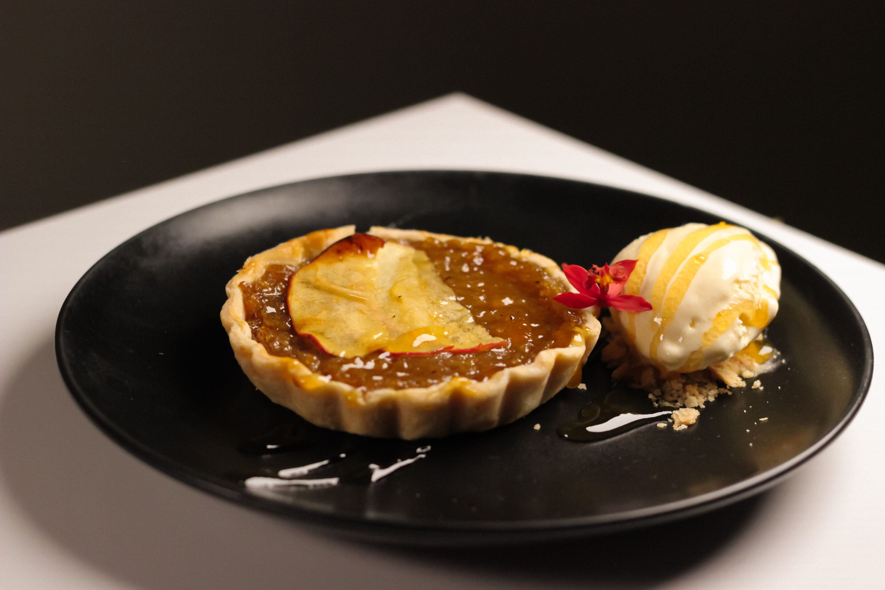 A small tart with caramelized apple slices is served on a black plate with a scoop of vanilla ice cream drizzled with sauce and garnished with a red edible flower.