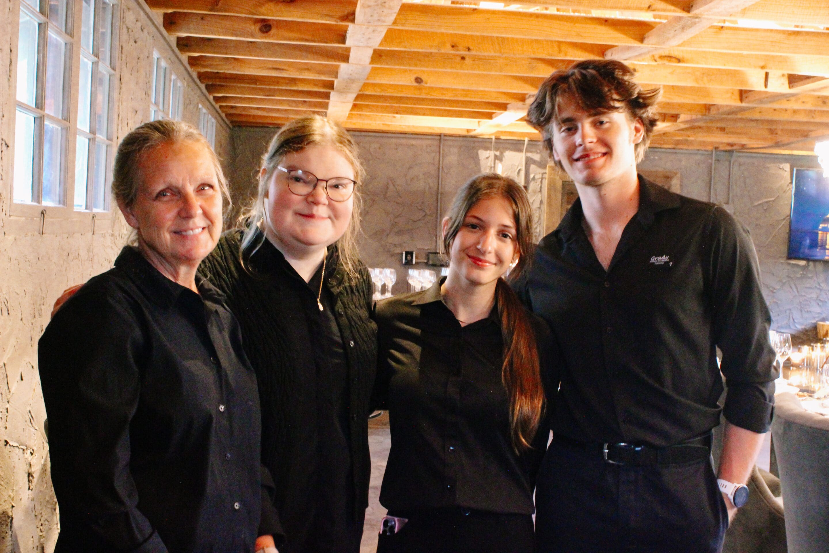 Four people stand together indoors, smiling at the camera. They are dressed in black shirts and appear to be in a rustic setting with exposed wooden beams and stone walls. The atmosphere looks friendly and warm.