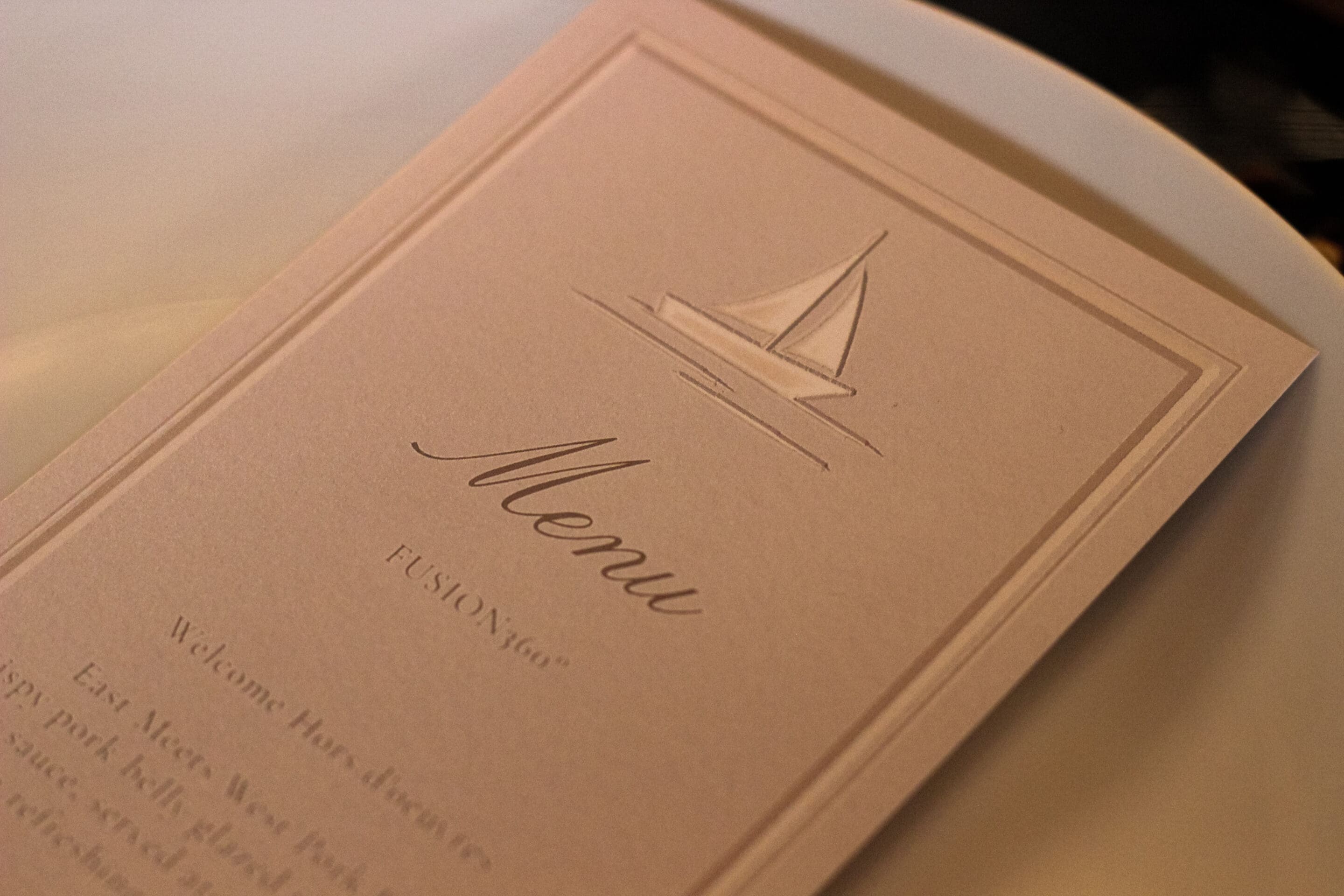 A close-up of an elegant, light-colored menu card with a simple sailboat illustration at the top, placed on a white plate. The word Menu is prominently displayed in cursive.