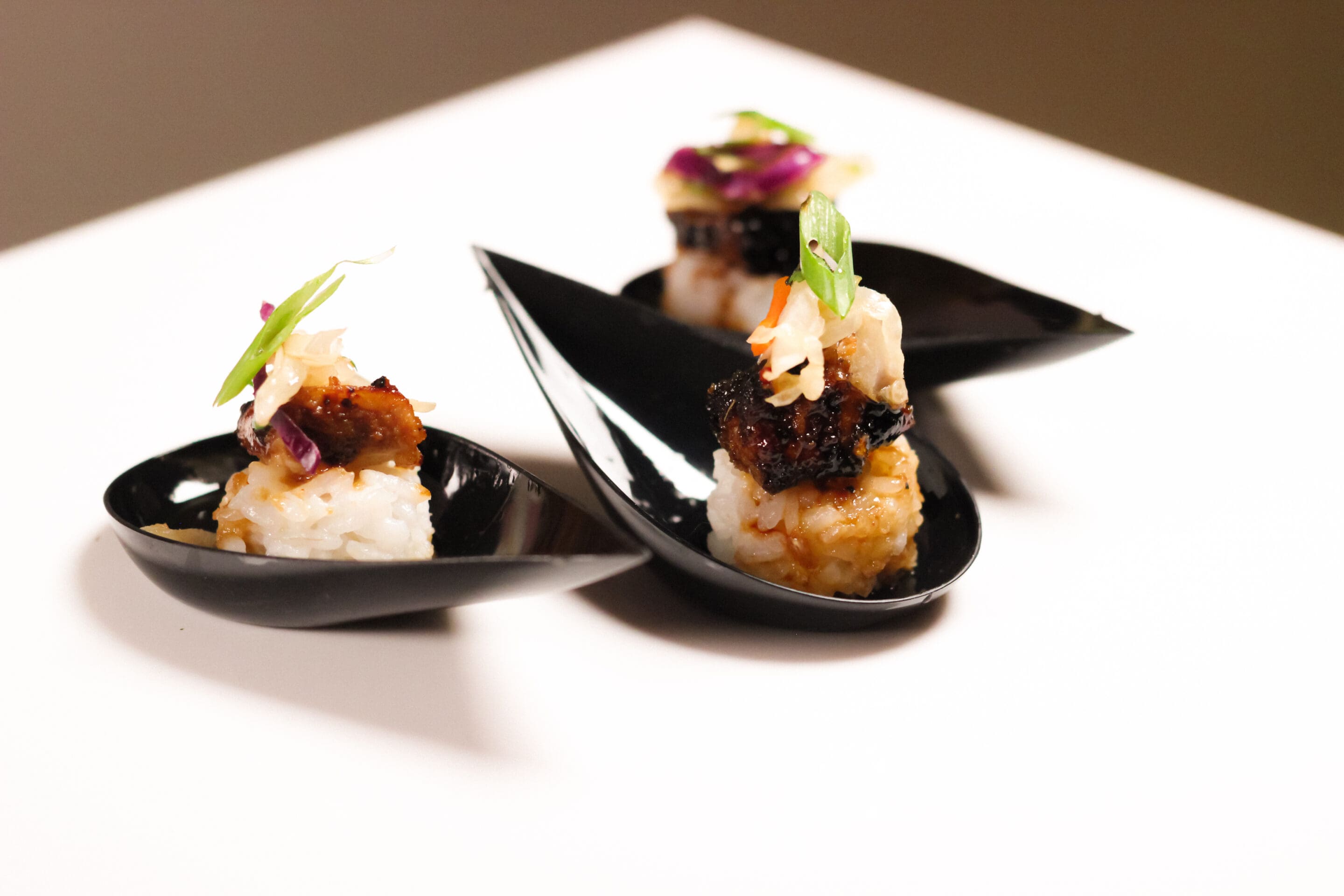 Three small servings of sushi rice topped with grilled meat, vegetables, and garnishes are presented on black, curved serving spoons against a white background.