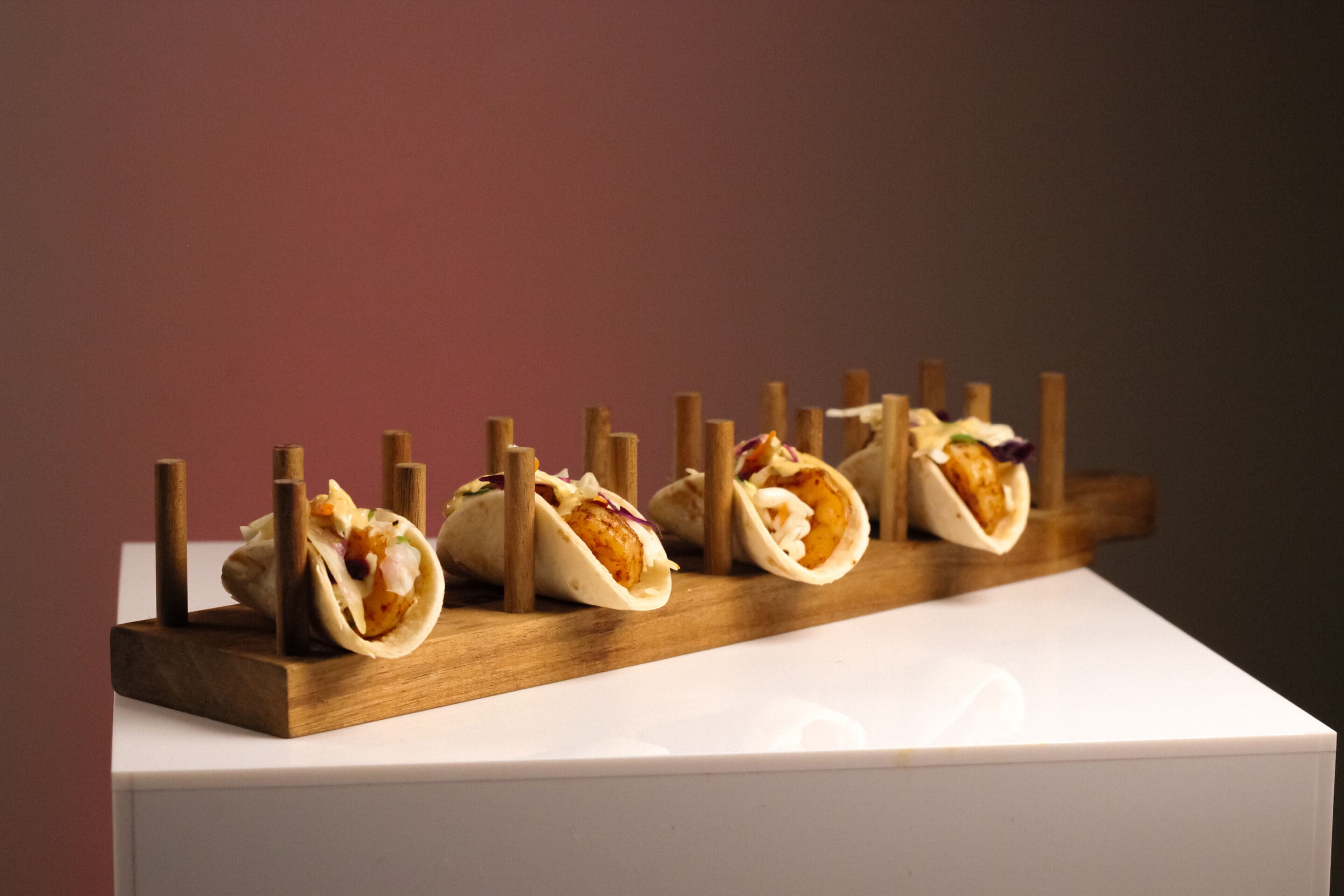 A row of small tacos filled with vegetables and meat is displayed on a wooden taco holder, set on a white surface against a gradient background.
