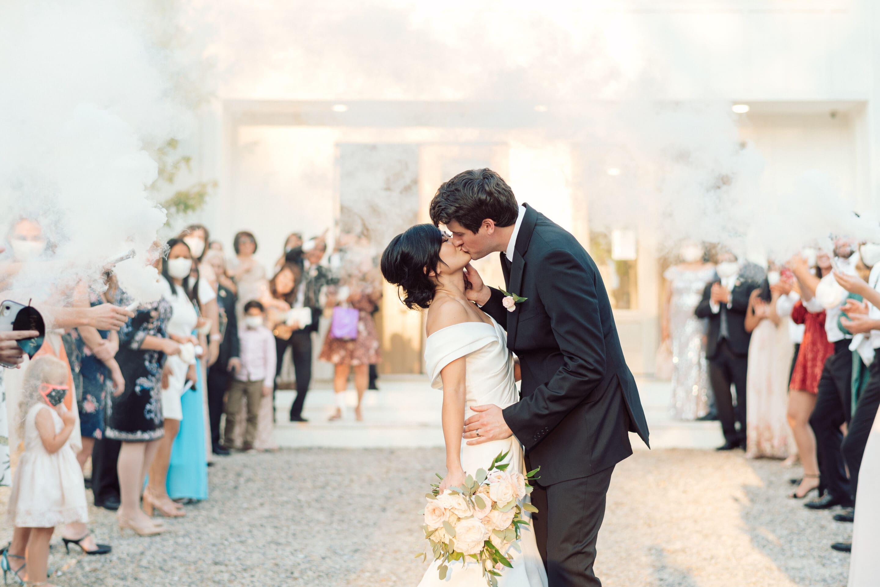 A bride and groom kiss outdoors as wedding guests celebrate around them with smoke bombs. The bride holds a bouquet, and guests in colorful attire cheer and take photos in the background.