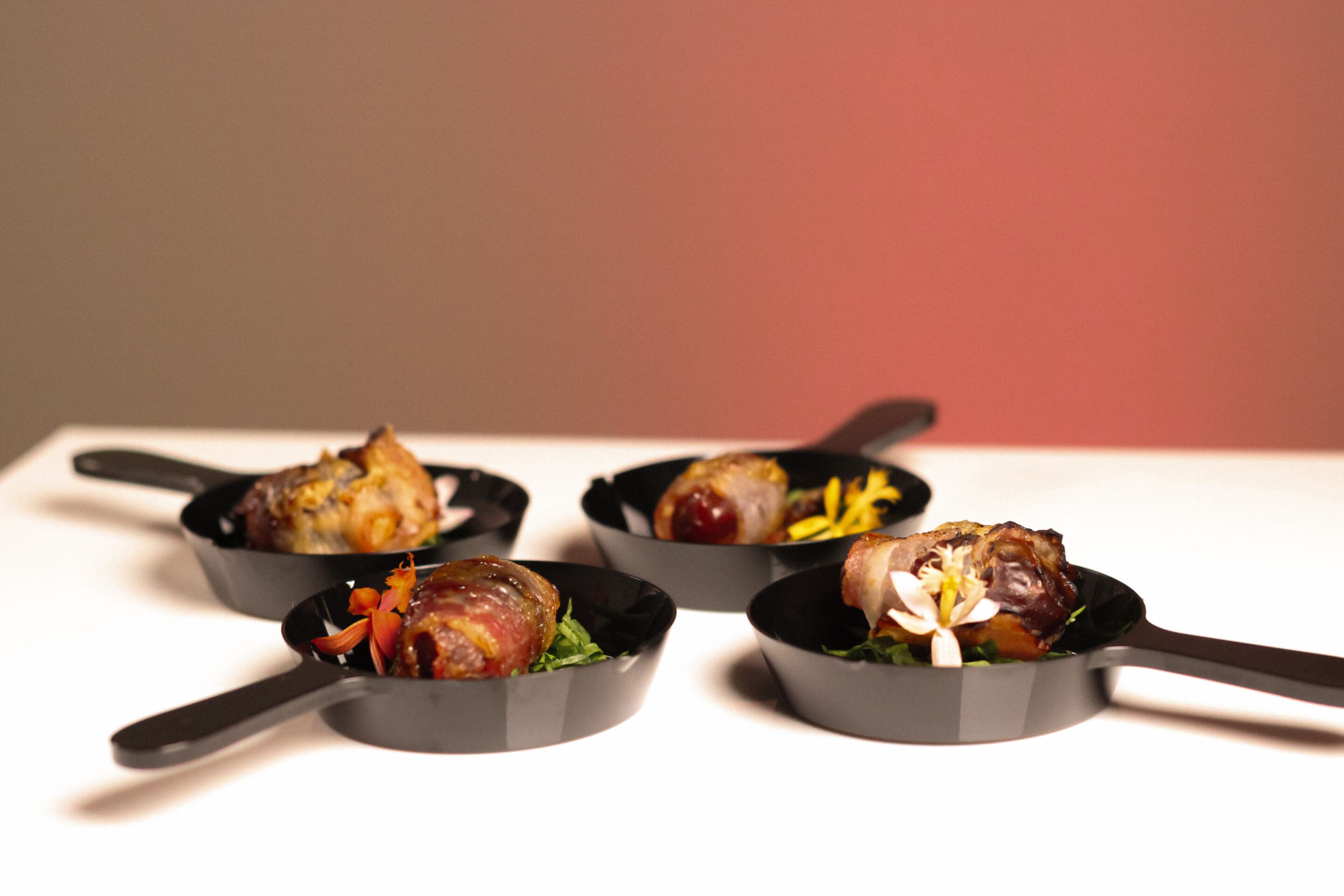 Four small black skillets, each holding a bite-sized appetizer garnished with colorful edible flowers and greens, are arranged on a white surface with a warm, softly lit background.