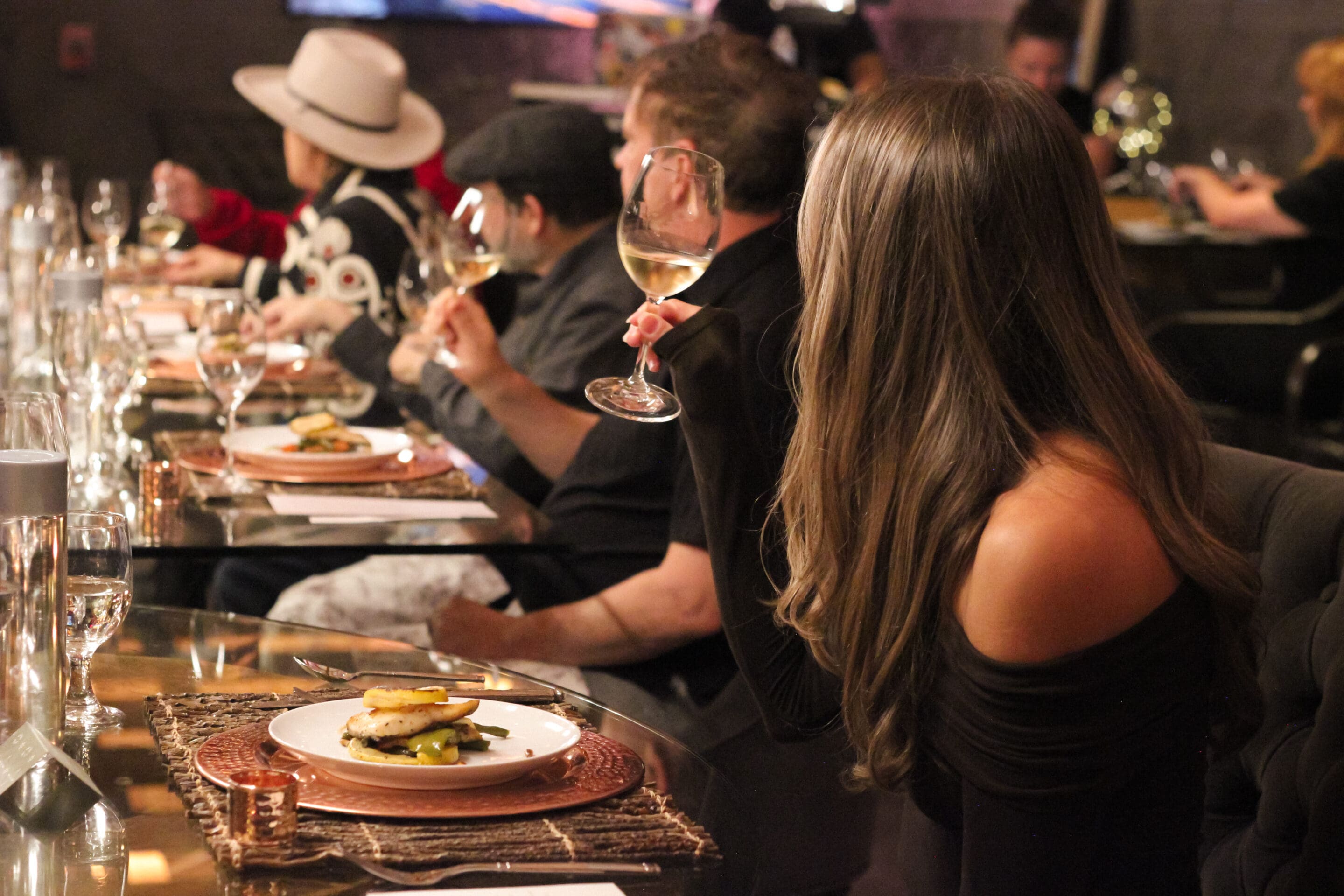 A group of people sit at a dining table, enjoying a meal. The focus is on a woman in a black off-shoulder dress holding a wine glass. Plates of food and glasses are set in front of each guest. The setting is warm and elegant.