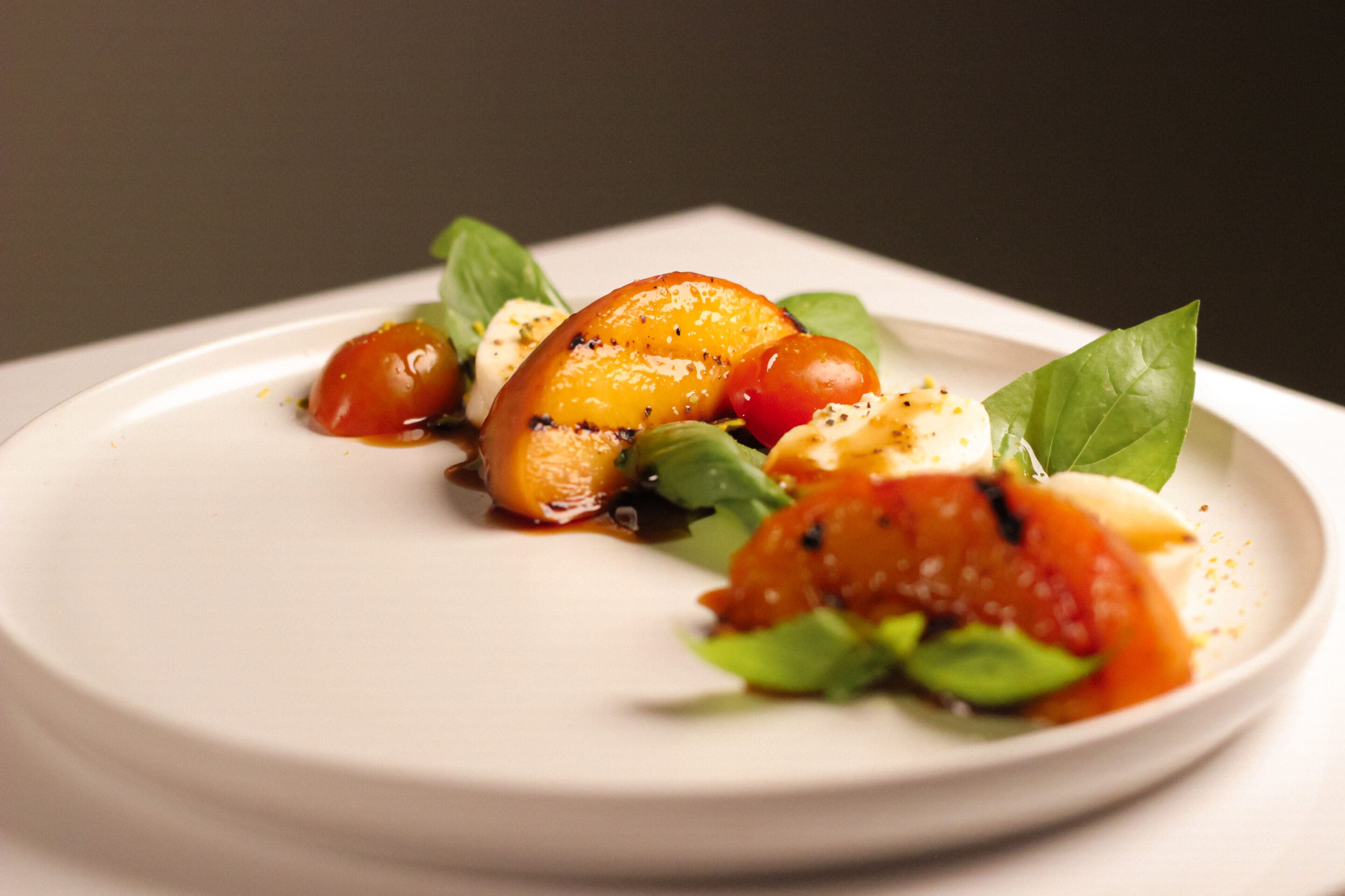 A white plate with a caprese salad, featuring slices of tomato, mozzarella, fresh basil leaves, and a drizzle of balsamic glaze, arranged artfully on the plate.