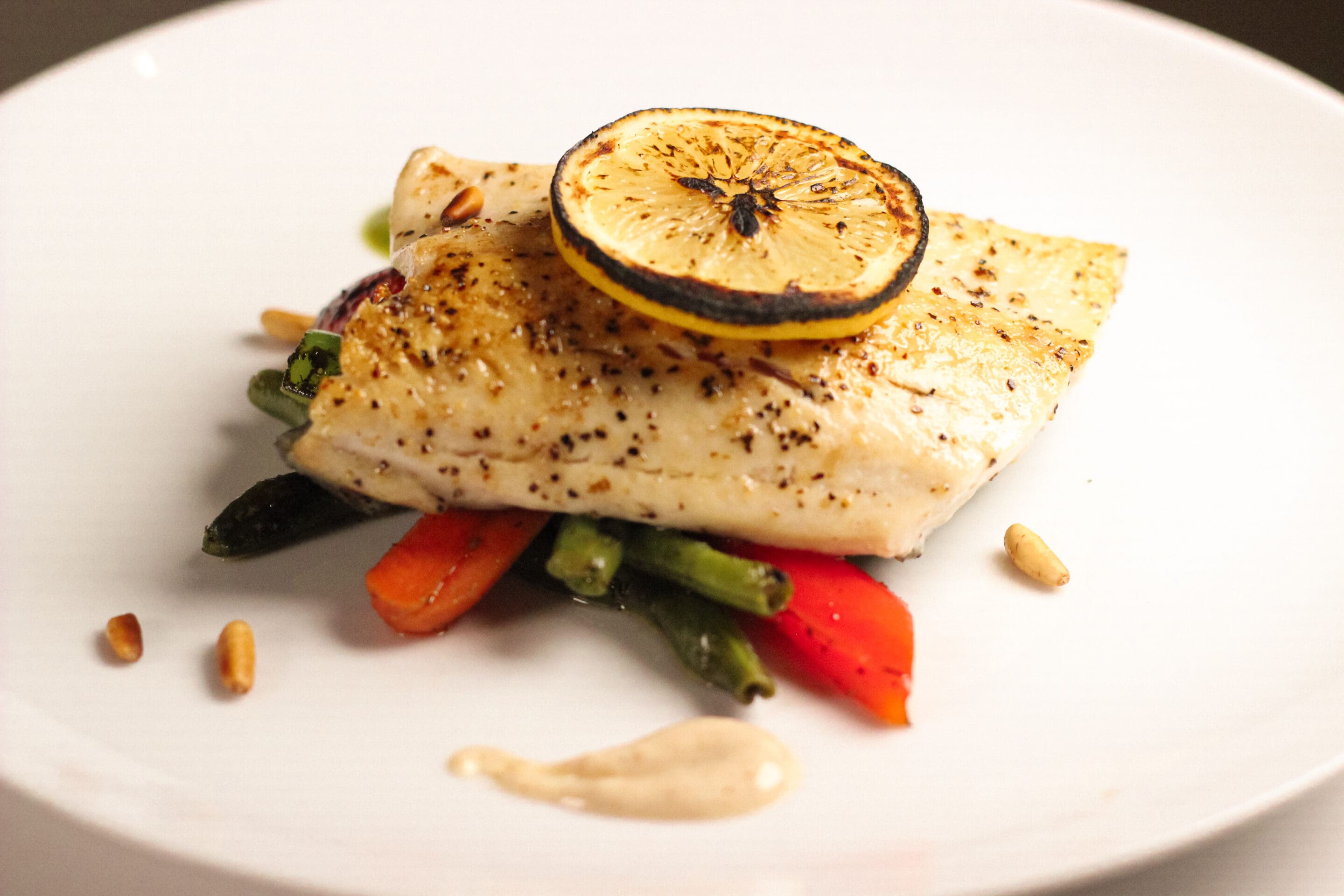 A fillet of grilled fish topped with a slice of roasted lemon, served over sautéed mixed vegetables, garnished with pine nuts and a dollop of creamy sauce on a white plate.