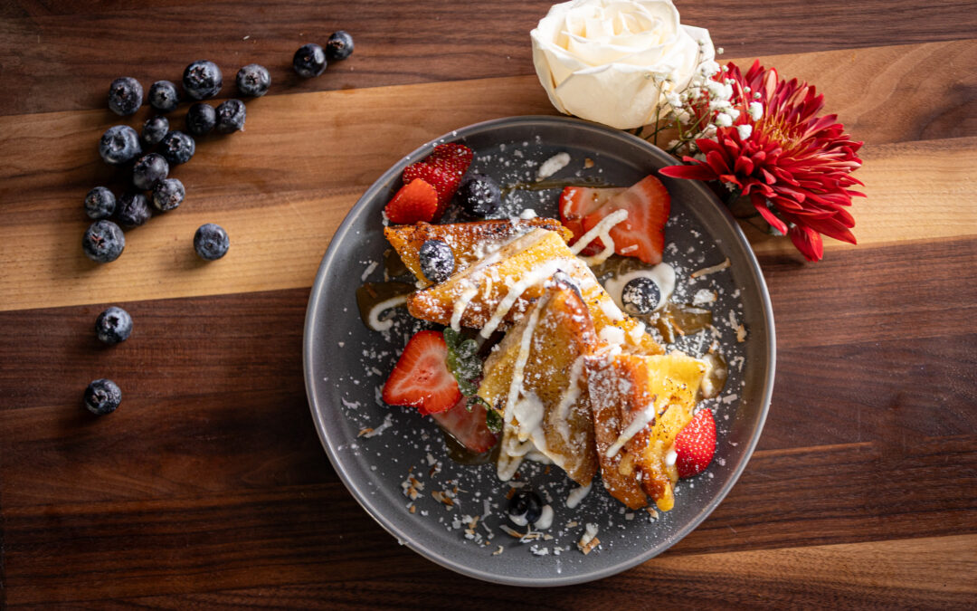 A plate of French toast topped with strawberries, blueberries, cream, and powdered sugar sits on a wooden table with scattered blueberries and two flowers—one white and one red—next to the plate.
