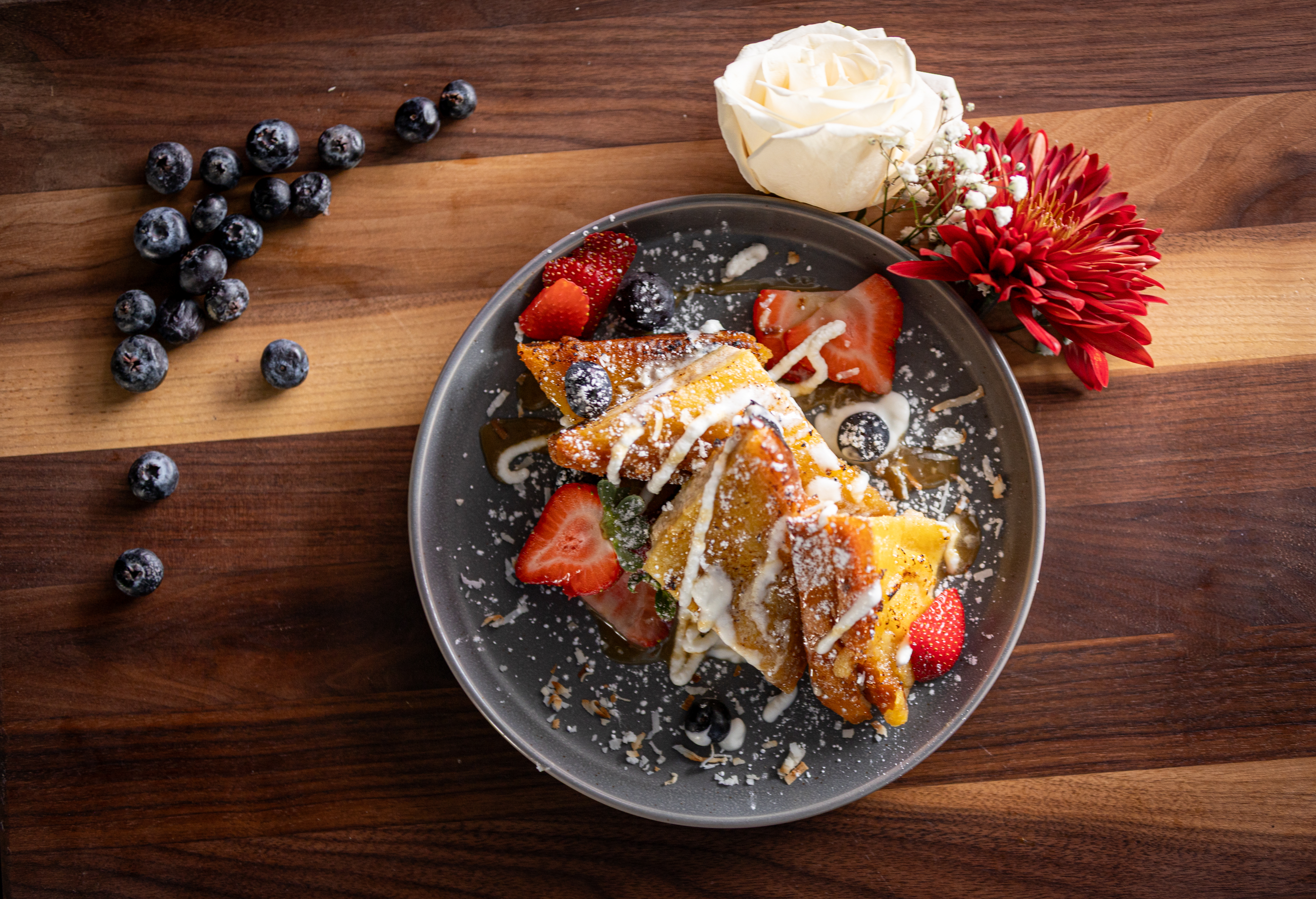 A plate of French toast topped with strawberries, blueberries, cream, and powdered sugar sits on a wooden table with scattered blueberries and two flowers—one white and one red—next to the plate.