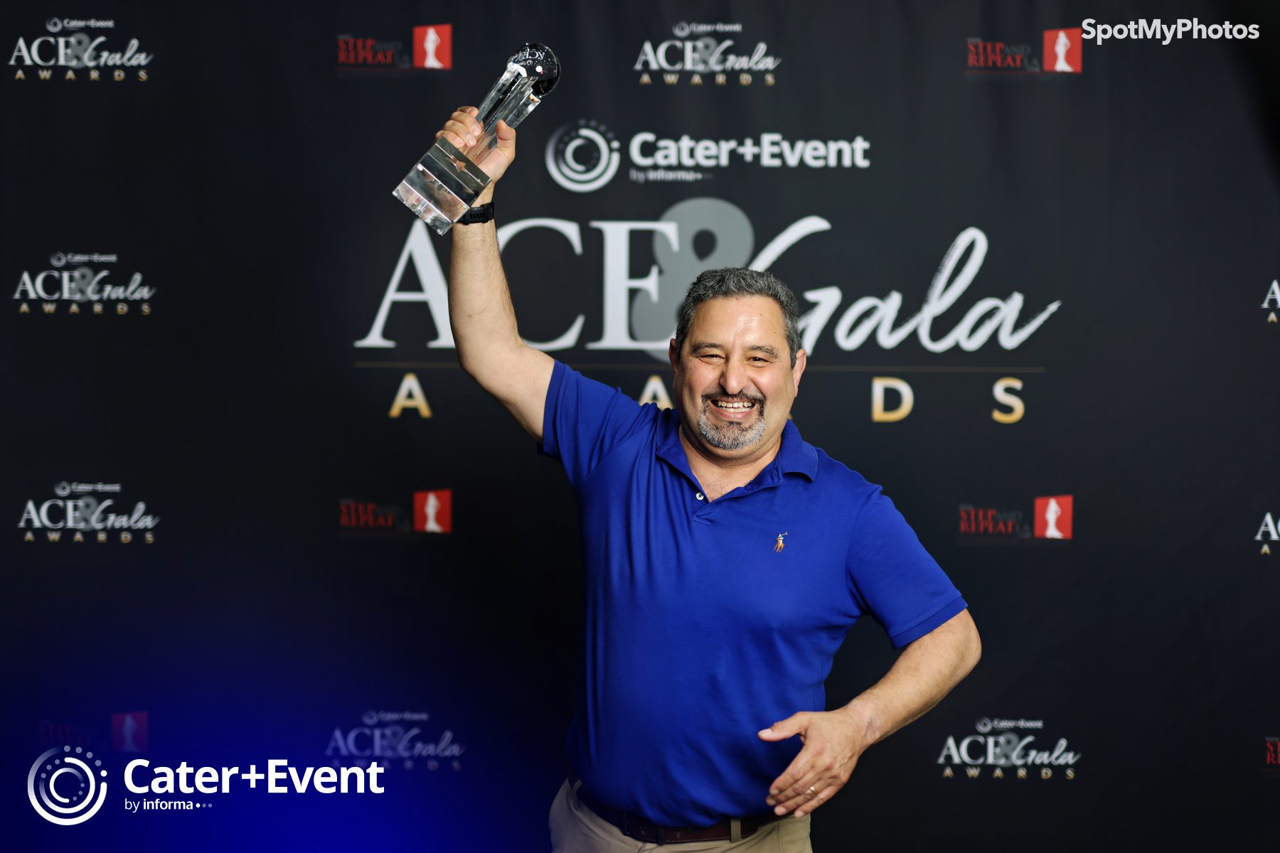 A man in a blue polo shirt smiles and raises a glass award in one hand while posing in front of an ACE Gala Awards step-and-repeat backdrop.