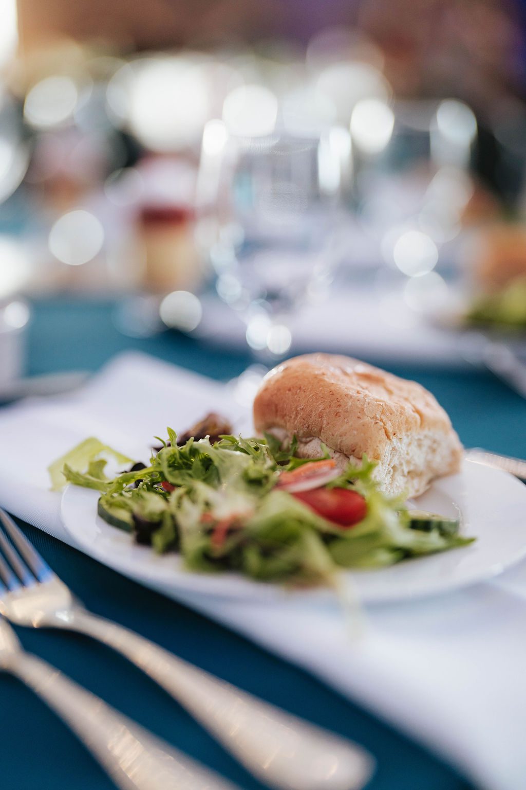 A close-up of a plate with a leafy green salad and a bread roll set on a table with a white napkin, silverware, and blurred glassware in the background.