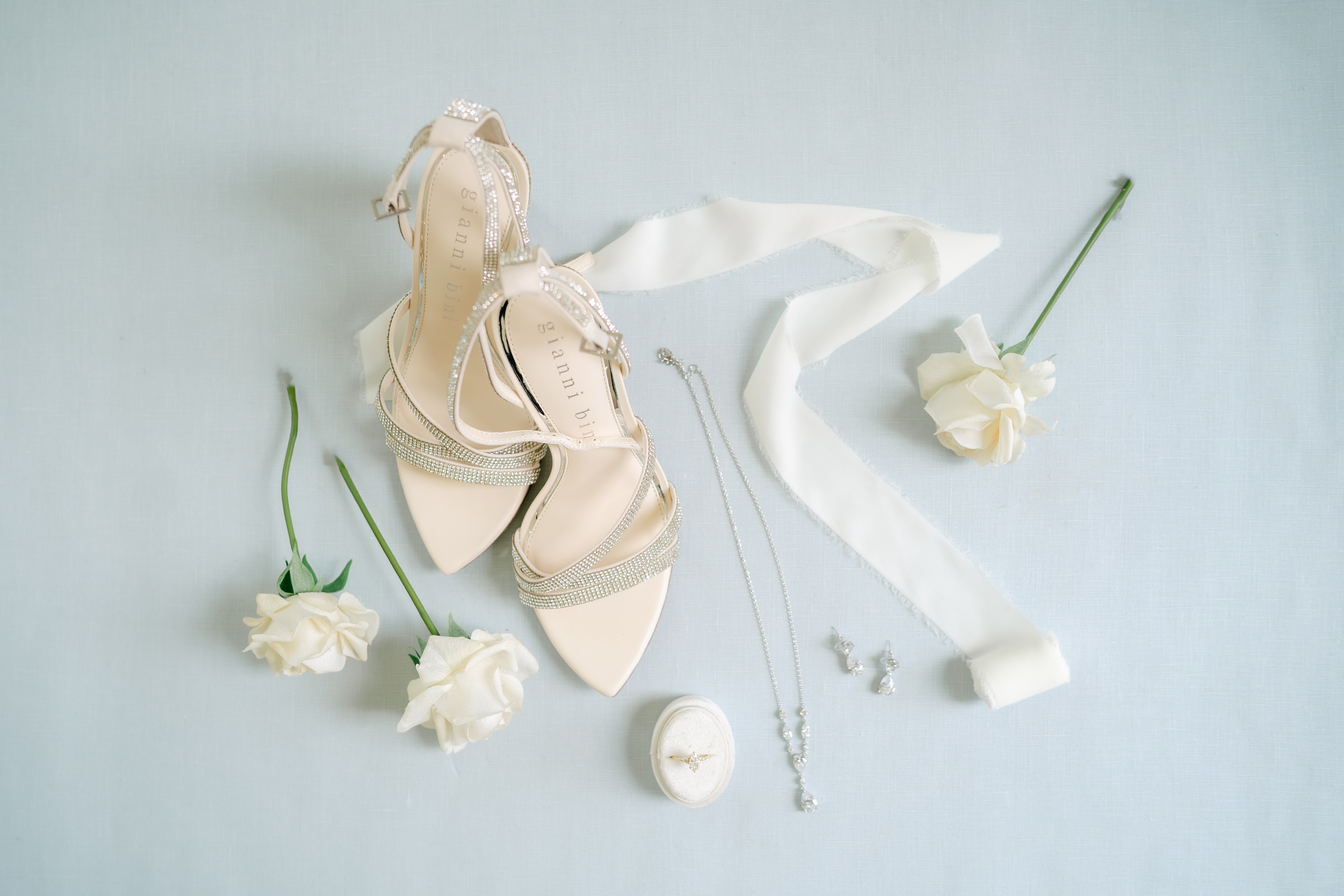 Elegant bridal accessories including beige high-heeled sandals, a necklace, earrings, a ring in a box, white ribbon, and three white roses arranged on a light surface.