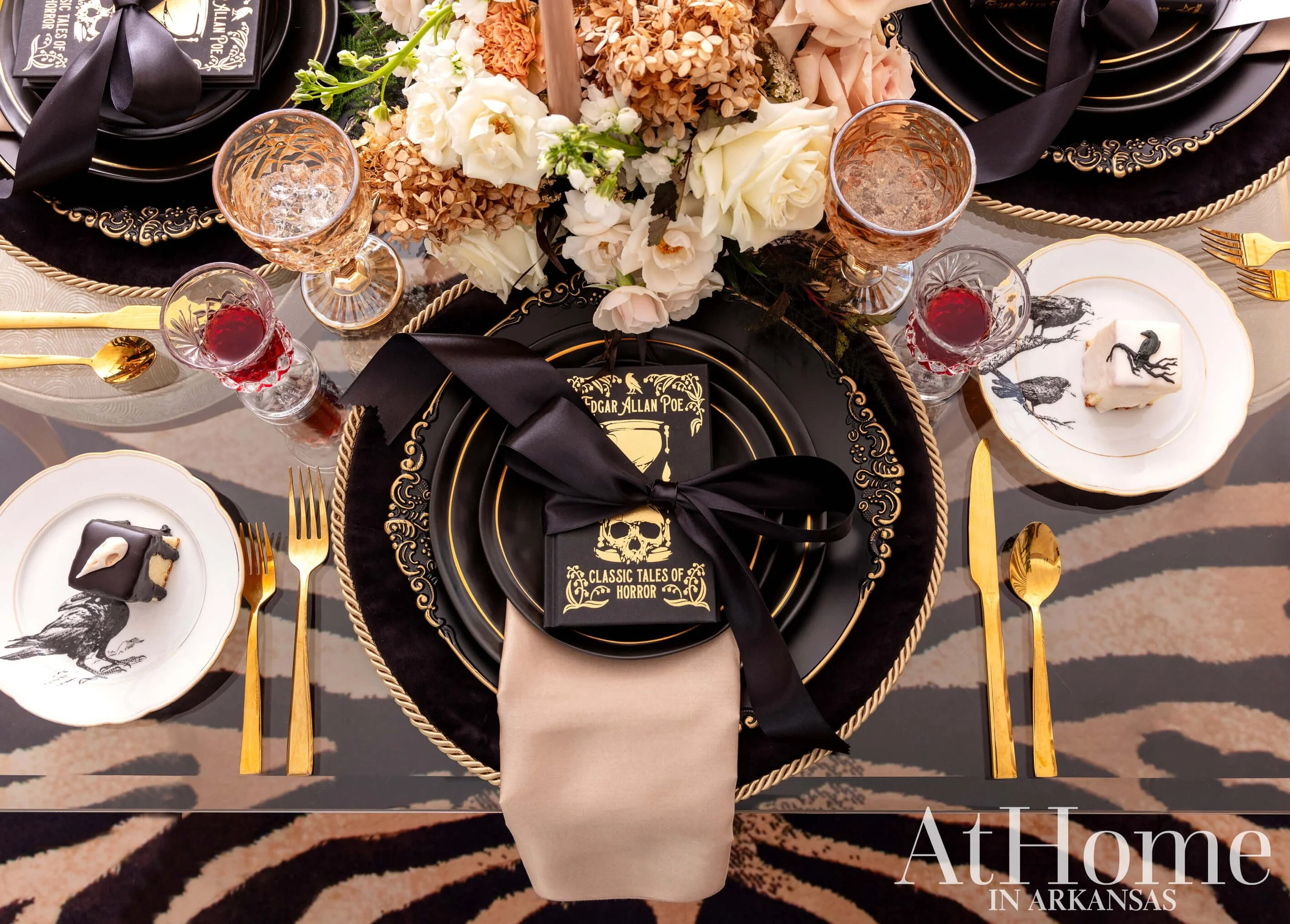 Elegant table setting with black and gold plates, gold cutlery, crystal glasses, a beige napkin, black ribbon, floral centerpiece, bird illustrations on plates, and At Home in Arkansas text in the bottom corner.