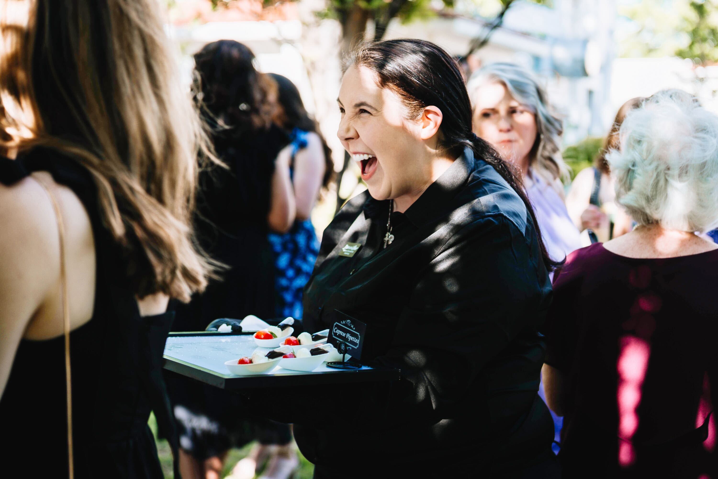 A woman in black, holding a tray of appetizers, laughs joyfully at an outdoor gathering with other people in the background. Sunlight filters through the trees above.