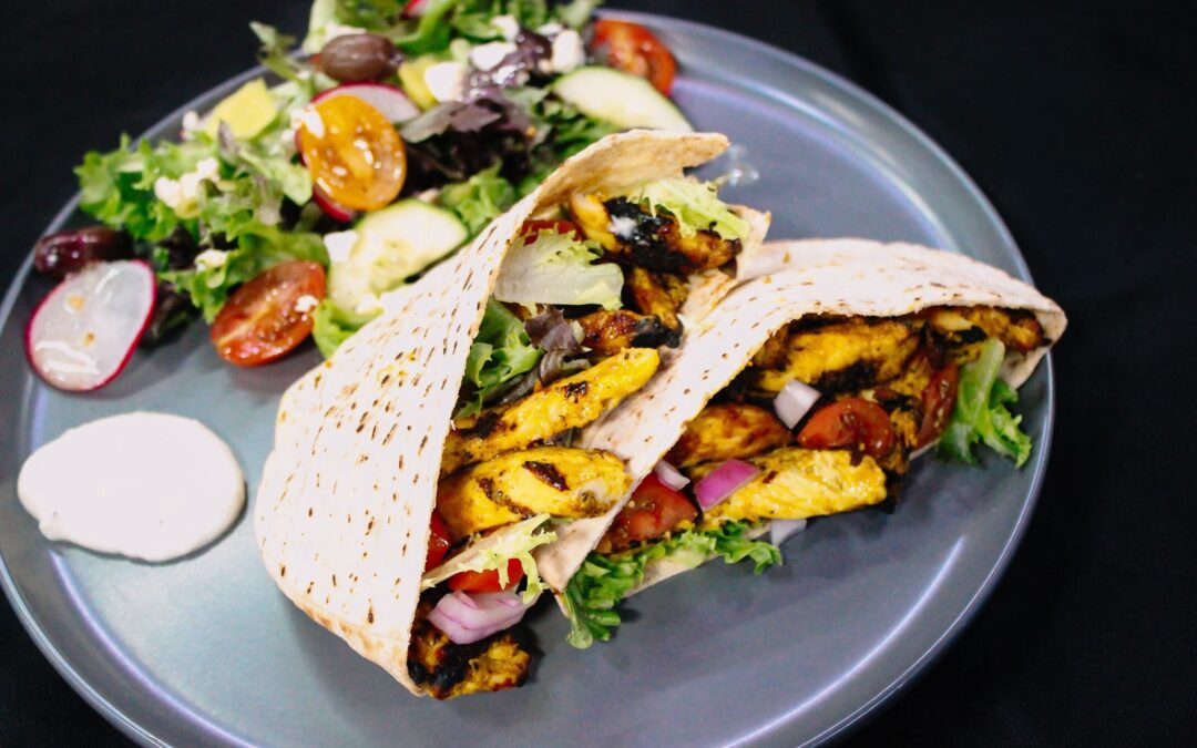 A plate with two pita pockets filled with grilled chicken, lettuce, tomatoes, and onions, served alongside a fresh salad with cucumbers, tomatoes, olives, and a dollop of white sauce on the side.