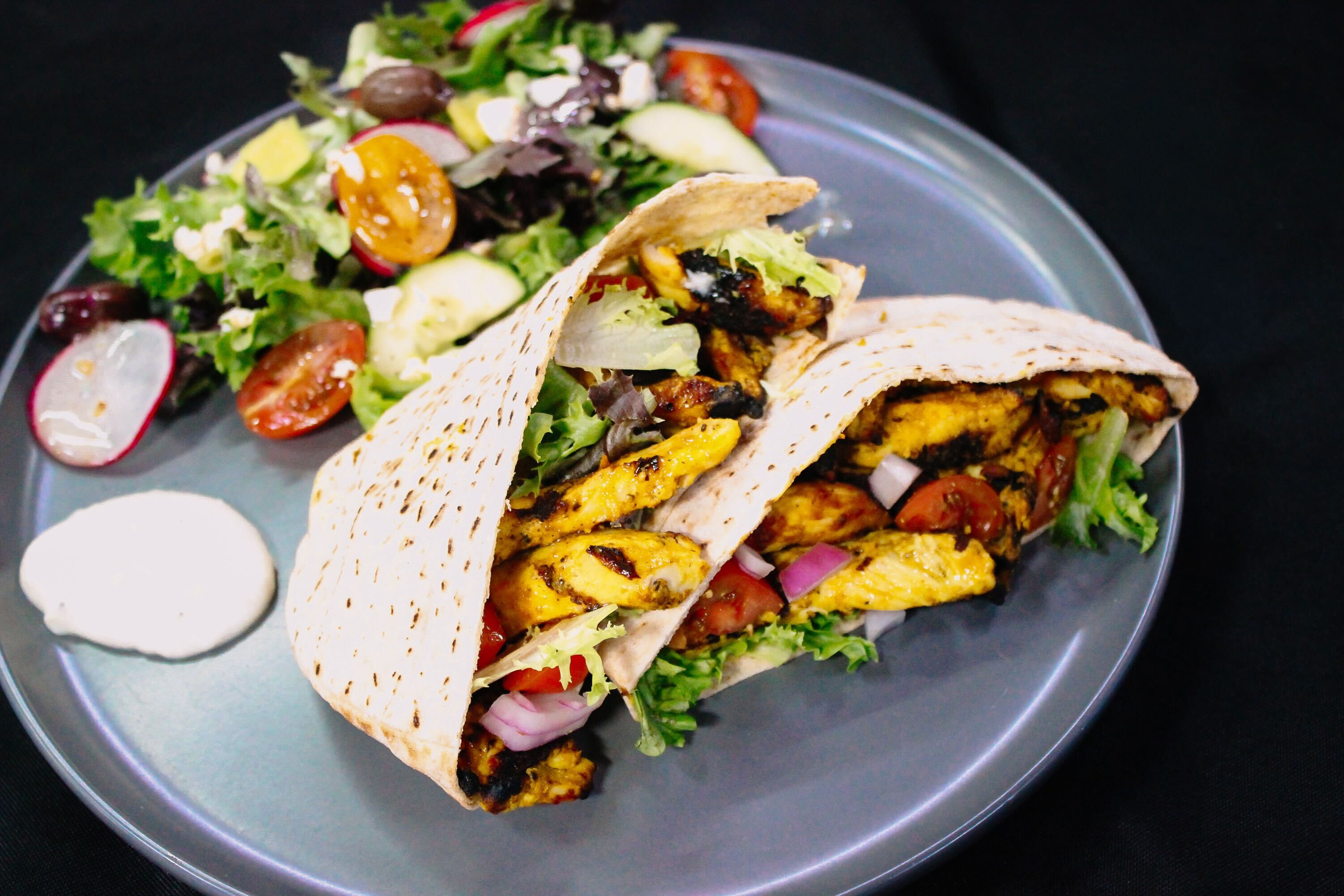 A plate with two pita pockets filled with grilled chicken, lettuce, tomatoes, and onions, served alongside a fresh salad with cucumbers, tomatoes, olives, and a dollop of white sauce on the side.