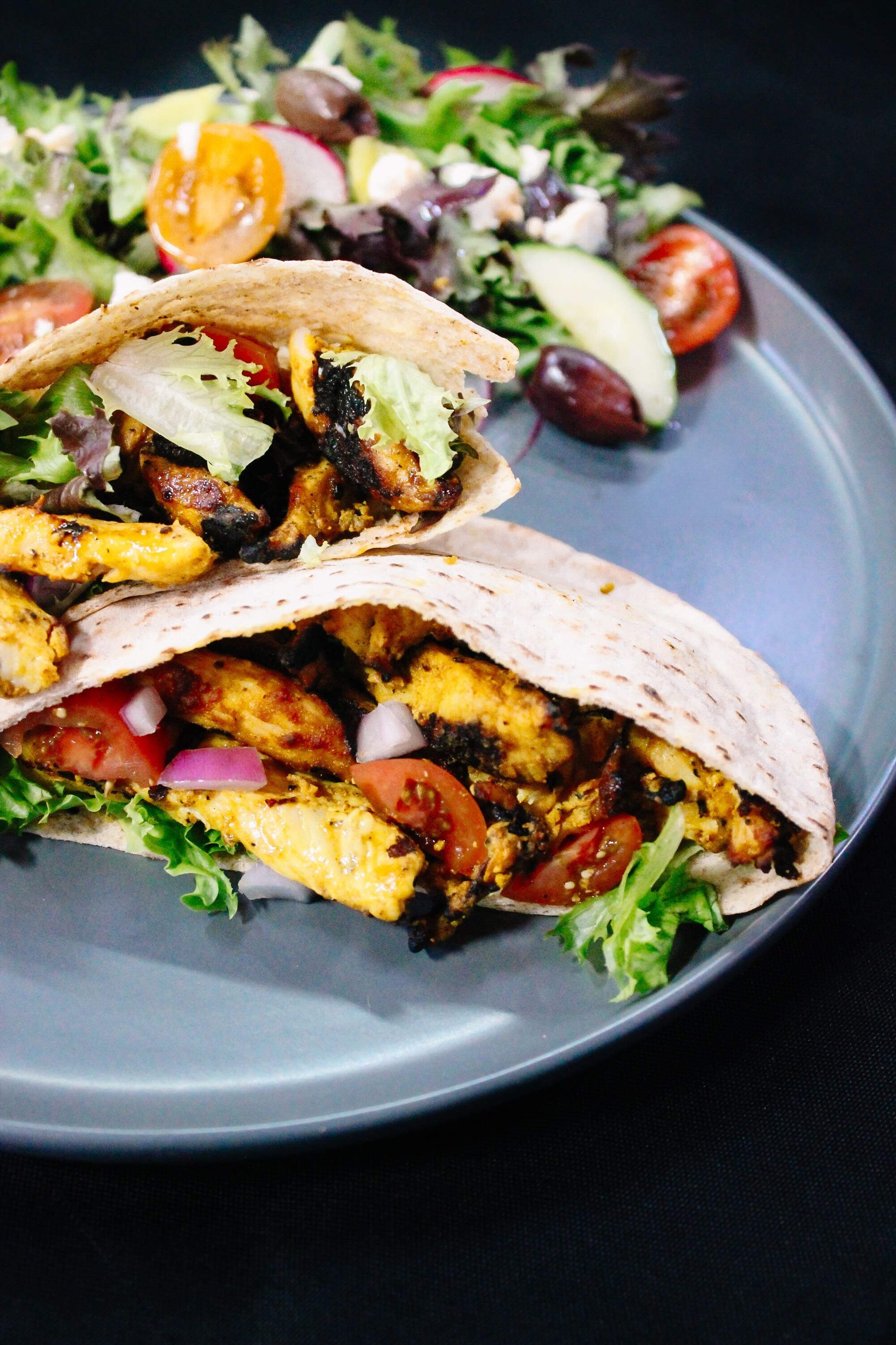 Two tacos filled with grilled chicken, lettuce, tomatoes, and onions on a gray plate, served with a side salad of mixed greens, tomatoes, cucumbers, and olives, against a black background.