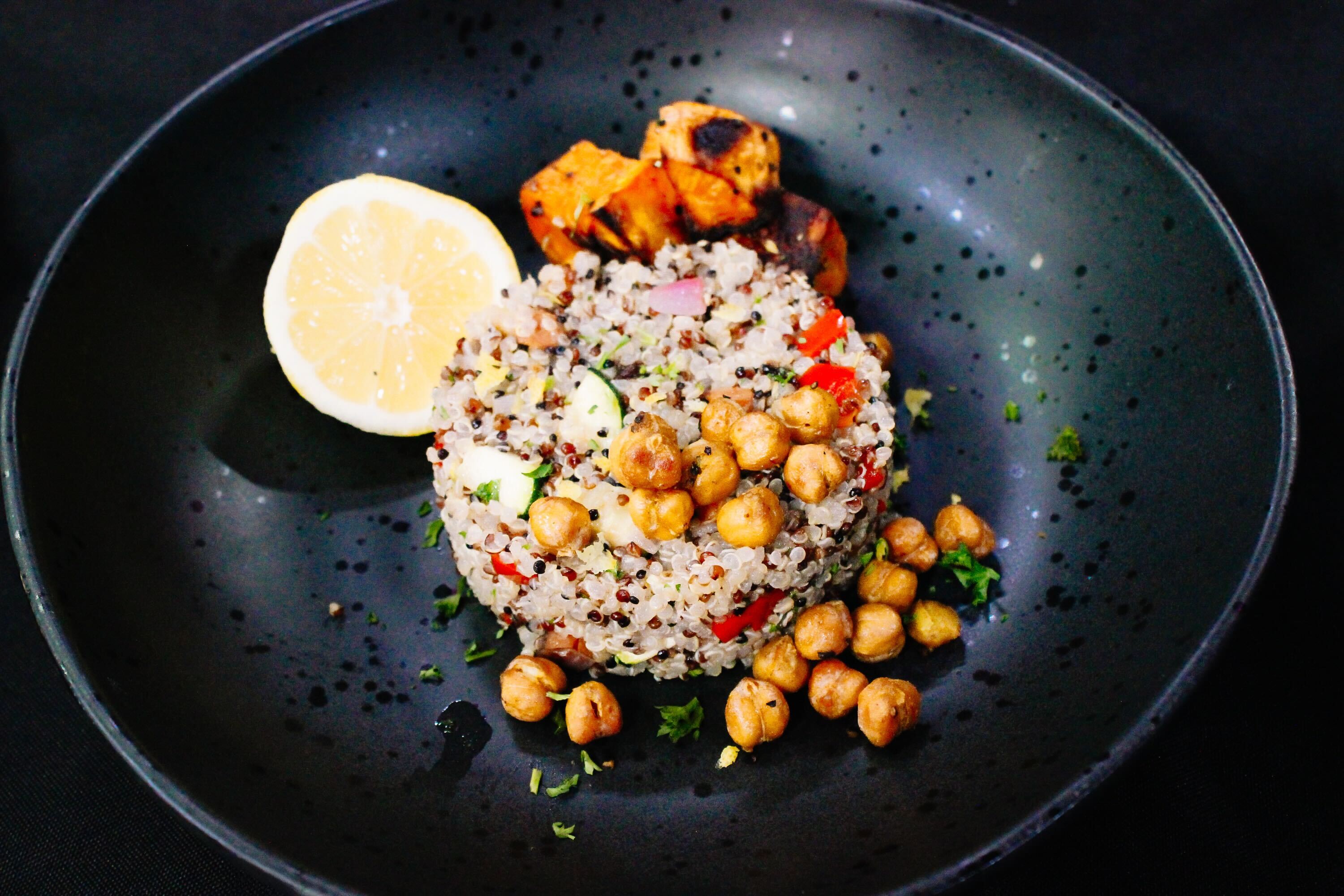 A black plate with a serving of quinoa salad topped with chickpeas, chopped vegetables, a lemon slice, and grilled cubes of tofu or paneer on the side, garnished with herbs.