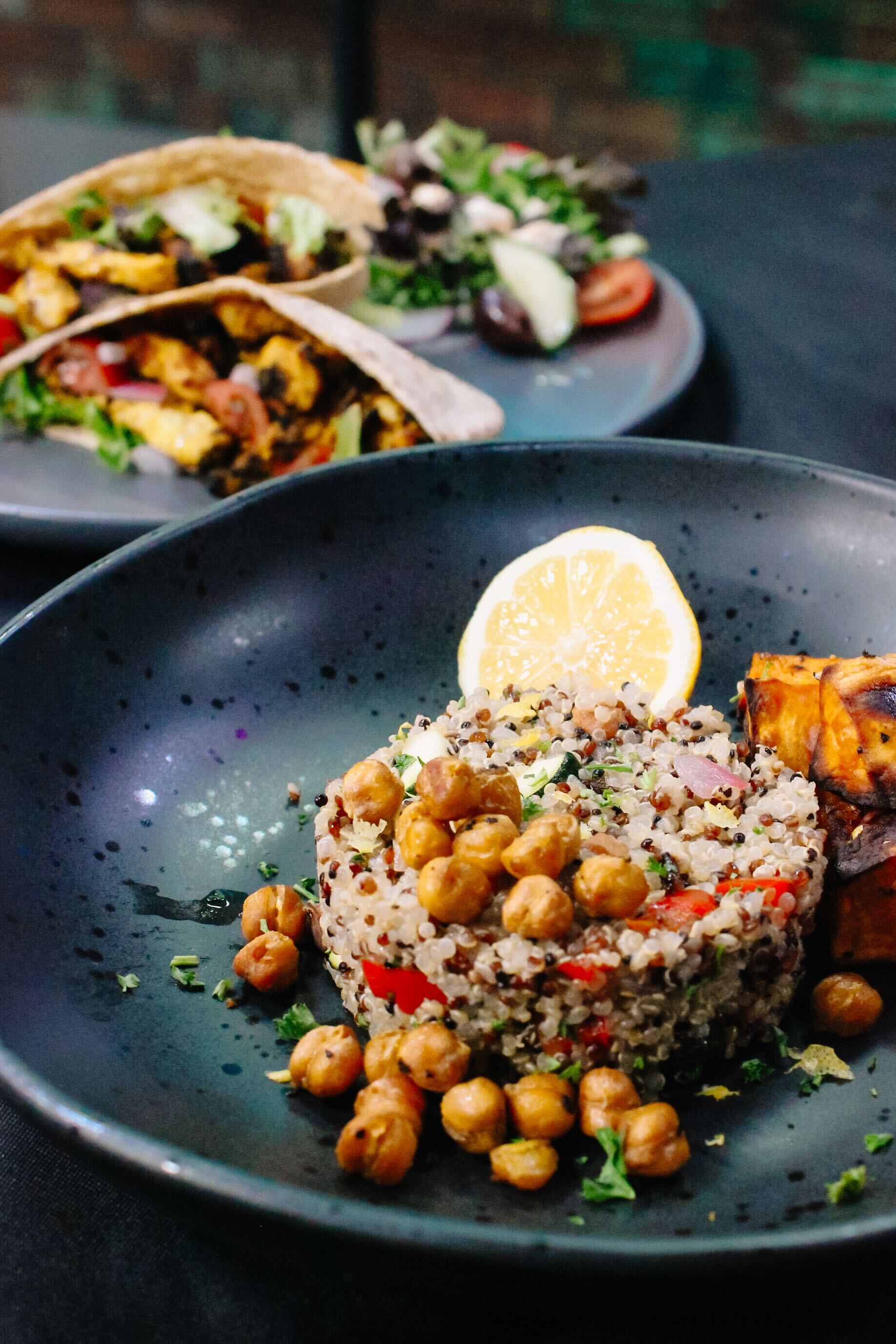 A black plate with quinoa salad topped with roasted chickpeas, a lemon wedge, and roasted sweet potatoes. In the background, pita wraps with vegetables and a small salad are visible on another plate.