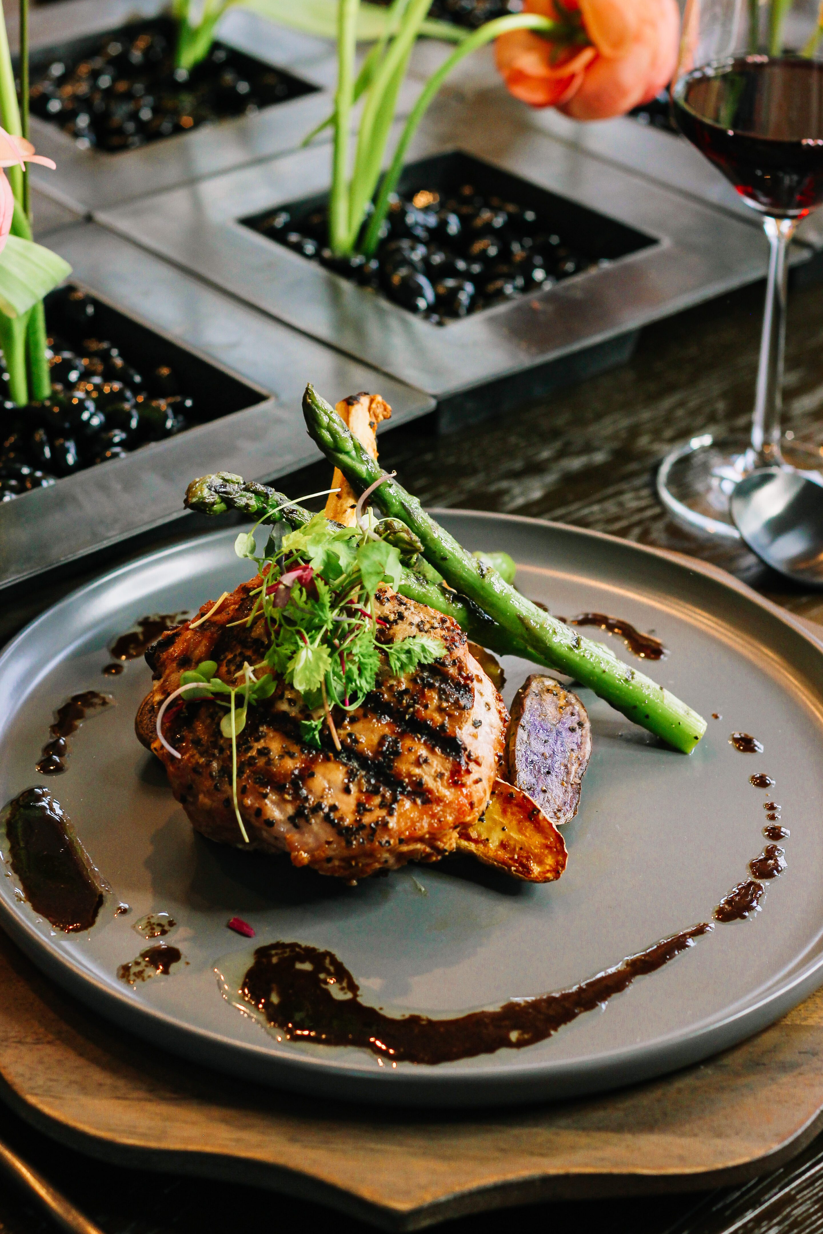 Grilled chicken breast served with asparagus, purple potato wedges, and microgreens, artfully plated with a dark sauce on a gray dish; a glass of red wine and decorative plants in the background.