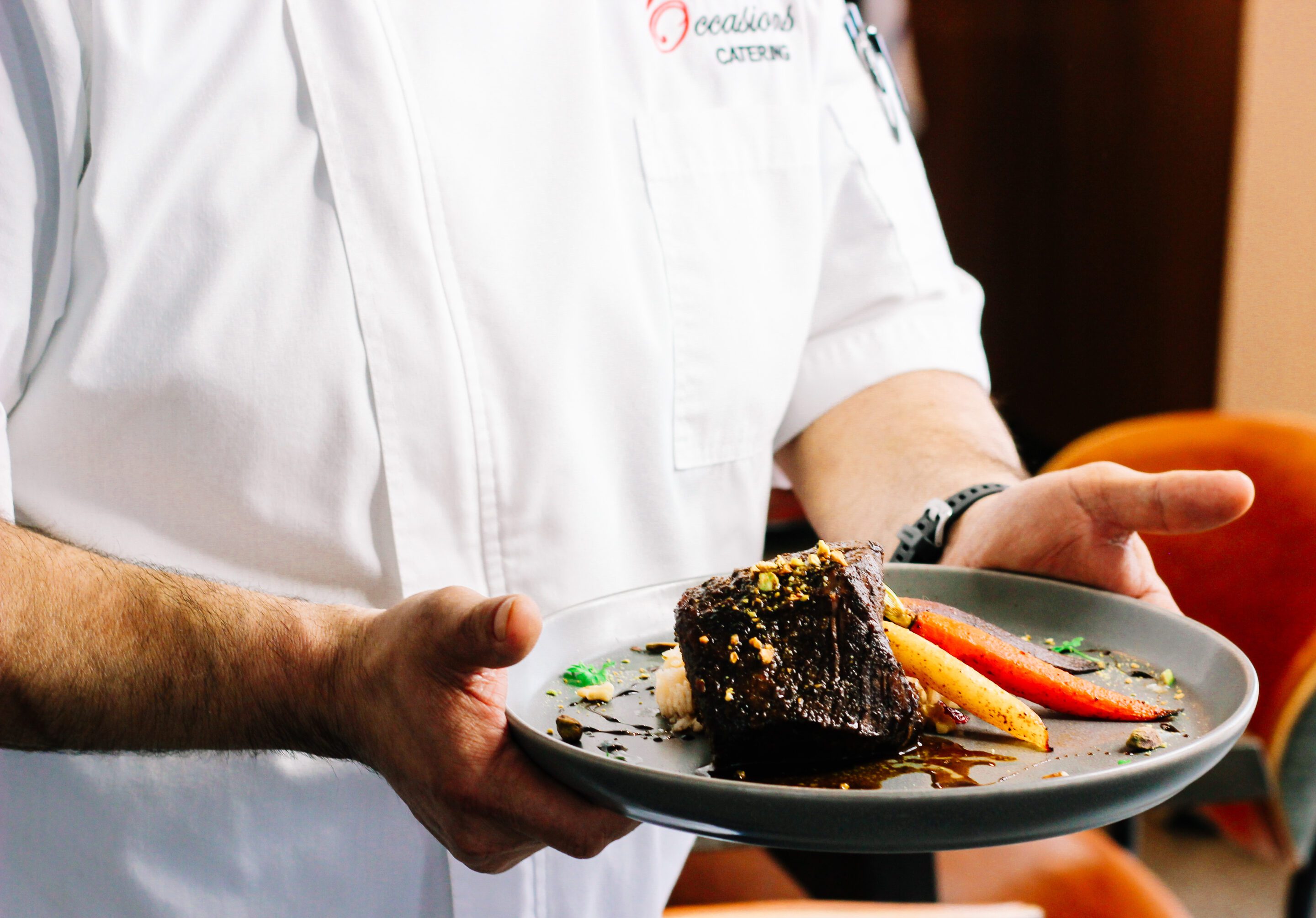 A chef in a white uniform holds a grey plate with a gourmet dish featuring a piece of braised meat, sauce, and colorful vegetables, including carrots, arranged artfully.