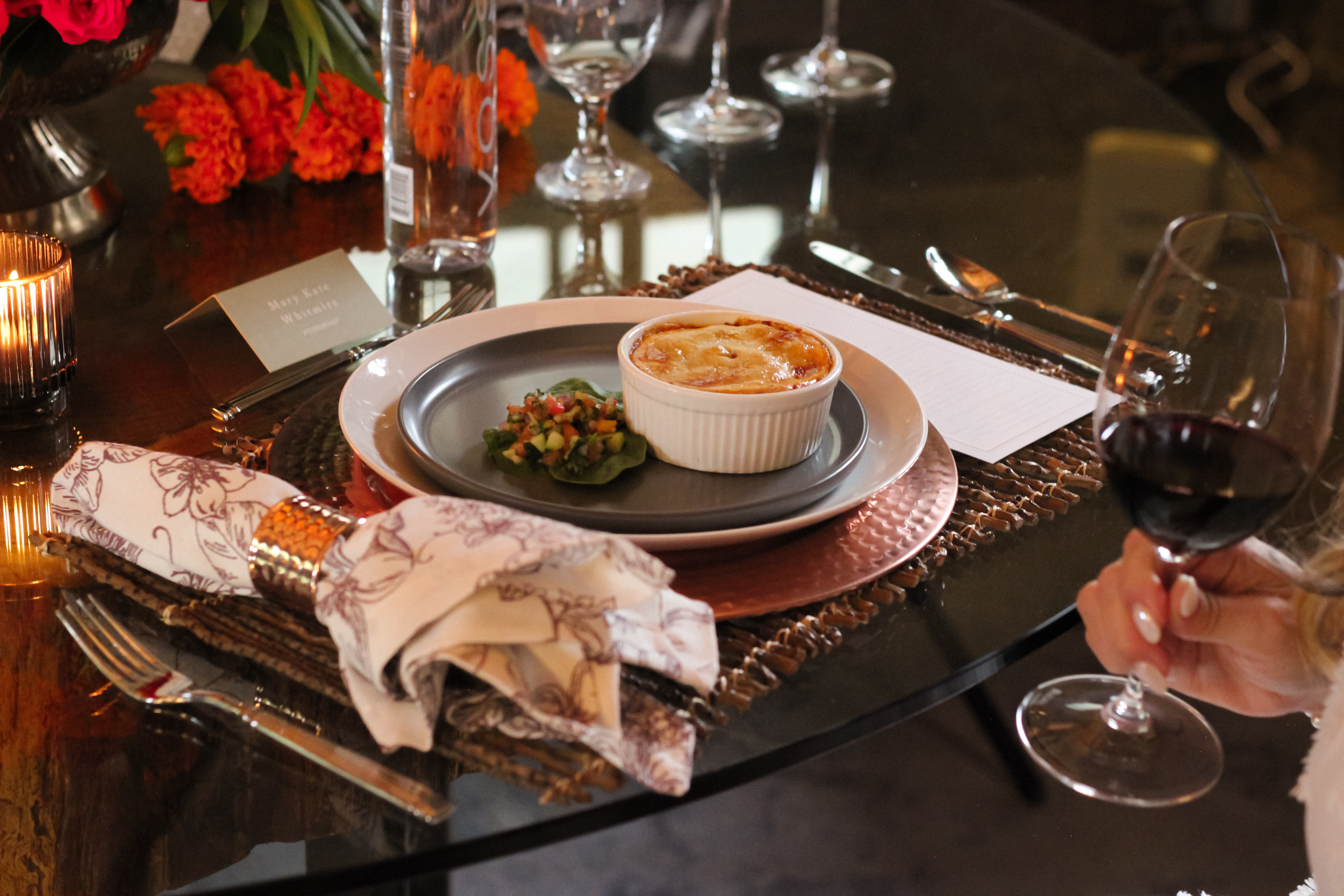 A table is set with a floral napkin, decorative napkin ring, and a plate holding a ramekin of baked dish and a small salad. A hand holds a glass of red wine. Bright flowers and place cards are in the background.