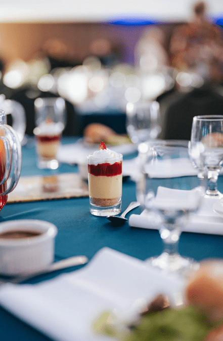 A small dessert in a glass with cream, fruit sauce, and a crumb base sits on a set table with glassware, plates, and napkins, creating an elegant dining atmosphere.