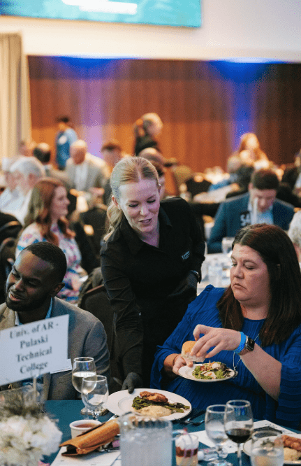 A server hands a plate of food to a seated woman at a formal event. Other attendees are visible at round tables in the background, and a sign on the table reads Univ. of AR-Pulaski Technical College.