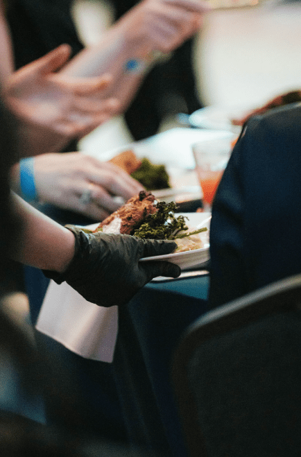 A server wearing a black glove holds a plate of food with greens and meat, while people sit at a table eating and conversing. The setting appears to be a formal event or dinner.