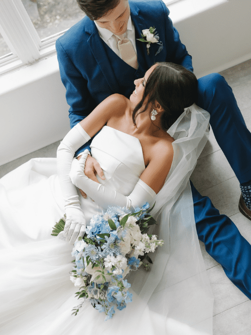 Southern Blue and White Wedding A bride in a white dress and veil lies in the arms of a groom in a blue suit. She holds a bouquet of white and blue flowers, and they gaze lovingly at each other near a window.