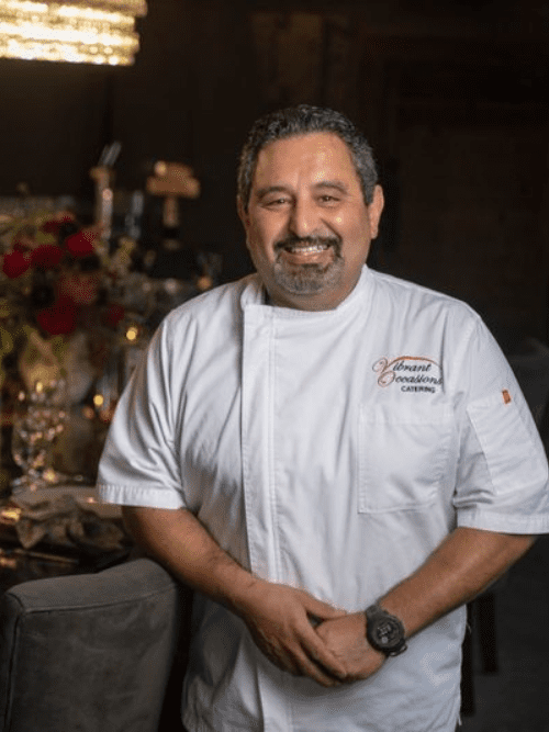 International Culinary Experience Supporting Local Foodbank A smiling chef with dark hair and a beard stands indoors, wearing a white chefs coat with Vibrant Occasions Catering embroidered on it. The background features a set dining table and soft lighting.