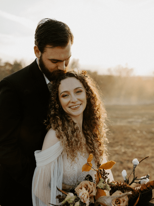 Whimsical Western Micro-Wedding A groom in a dark suit kisses the head of his smiling bride, who has curly hair and wears an off-the-shoulder white dress, holding a bouquet of autumn flowers outdoors at sunset.