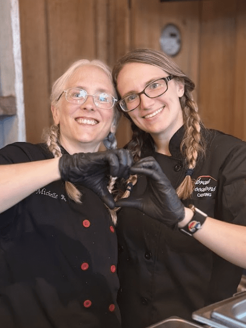 New Mission Statement Reflecting Heart-Centered Leadership Two women wearing glasses and black chef coats stand close together, smiling and making a heart shape with their gloved hands in a kitchen setting.