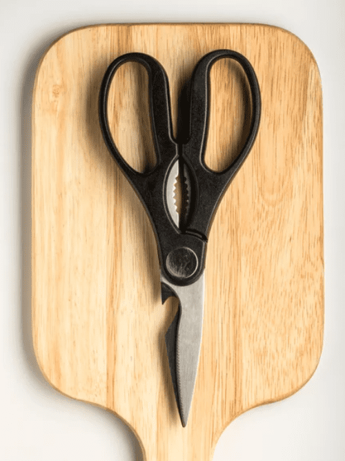 Improve Your Kitchen Shears A pair of black-handled kitchen scissors placed on a light wooden cutting board, viewed from above.