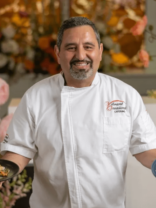 Corporate Culture Matters in Business A smiling chef in a white coat holds two plates of food, standing indoors with a blurred floral background. The coat reads Vibrant Occasions Catering on the chest.