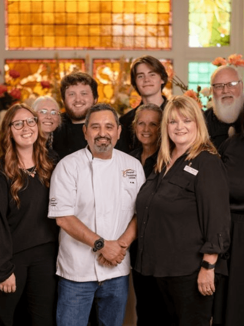 Best of 2025 Winner in 3 Categories A group of eight people, including a chef in a white coat, stand together and smile for the camera in front of colorful stained glass windows and floral decorations.