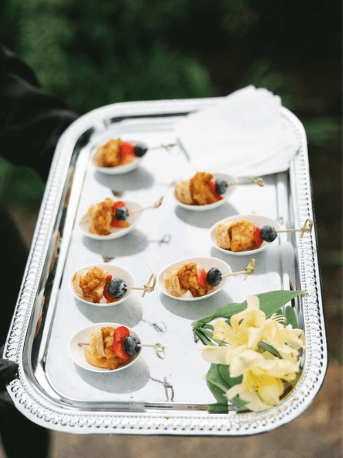 2026 Wedding Catering Trends Analysis A silver tray holds small white spoons with bite-sized appetizers skewered with blueberries and strawberries, alongside a napkin stack and yellow flowers, against a blurred green background.