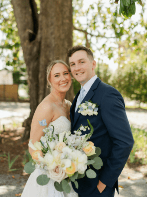 Lauren and Grant Posey A bride and groom smile while standing close together outdoors. The bride holds a large bouquet of white, yellow, and peach flowers. Both are dressed formally, with greenery and sunlight in the background.