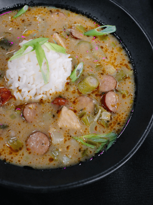 Community Spirit With Complimentary Gumbo at Saline County Fundraiser A bowl of creamy gumbo with sliced sausage, chopped vegetables, and a mound of white rice in the center, garnished with green onions, served in a black bowl on a dark background.