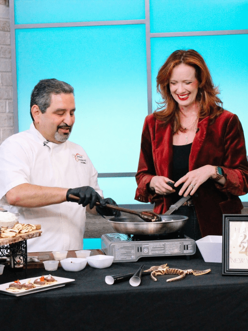 Elegant Winter Party Appetizers A chef in a white coat and black gloves cooks food in a pan while a smiling woman in a red blazer watches. They stand behind a table with plates, bowls, and cooking utensils in a brightly lit studio setting.