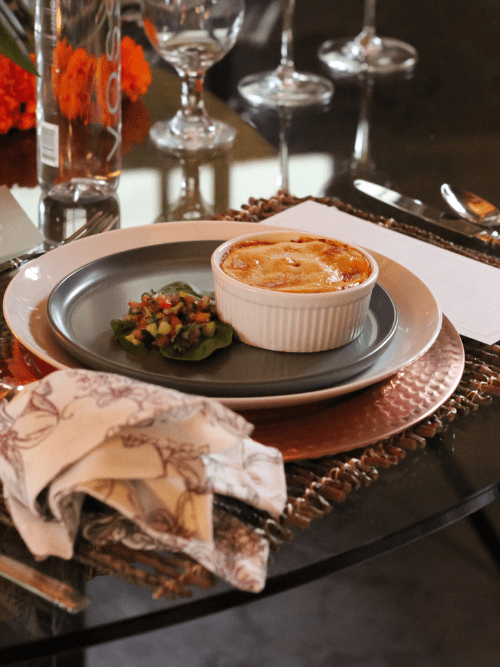 FUSION360 Culinary Experience A white ramekin with a baked dish and a small green salad sits on a gray plate, placed on a copper charger with a patterned napkin nearby on a set dining table.