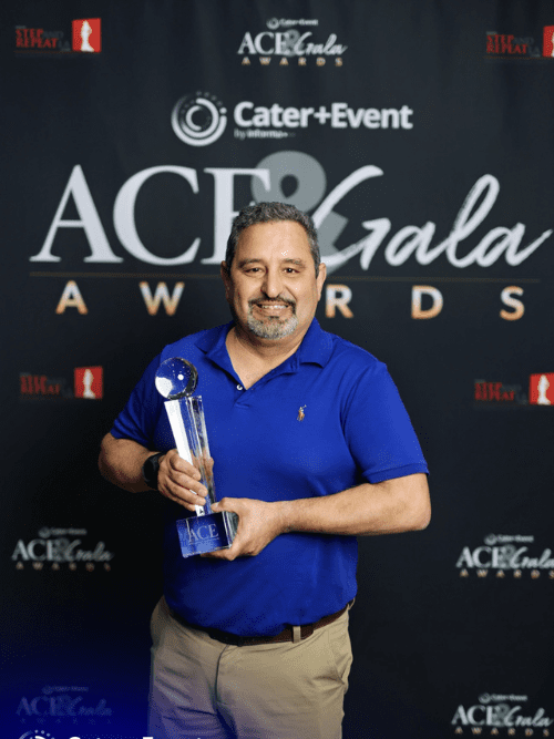 2026 Catered Micro Event of the Year A man in a blue shirt smiles while holding a glass trophy in front of a black backdrop with ACE Gala Awards and Cater+Event logos.
