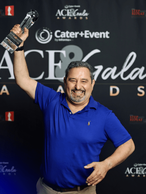 2026 Catered Micro Event of the Year A man in a blue polo shirt smiles and raises a clear award trophy in one hand against a black ACE Gala Awards backdrop.
