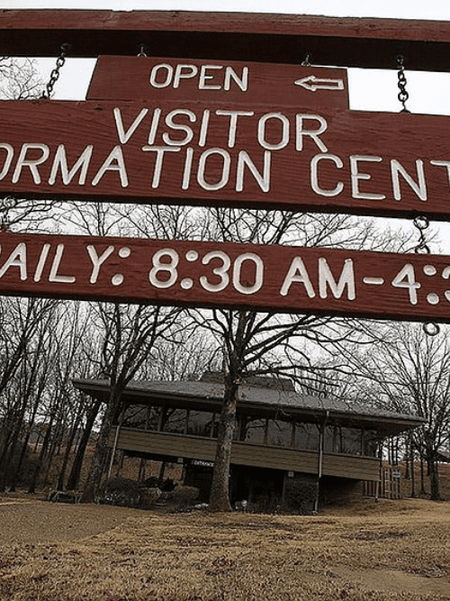 Dining news around Little Rock A wooden sign reads Open Visitor Information Center Daily: 8:30 AM - 4:30 PM with an arrow pointing left. Bare trees and a building are visible in the background on a cloudy day.