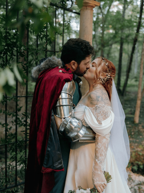 Whimsical Woodland Wedding A couple kisses outdoors; the groom wears medieval armor and a red cape, while the bride wears an off-shoulder wedding dress with lace sleeves and a veil, holding a bouquet in a forest setting.