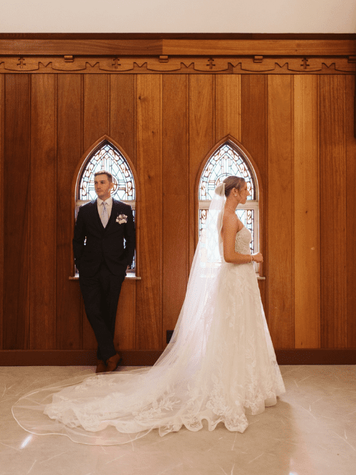 Classic Blue and White Wedding A bride in a white gown and veil stands facing a groom in a dark suit, both in front of wooden paneling with arched stained-glass windows, in a softly lit indoor setting.