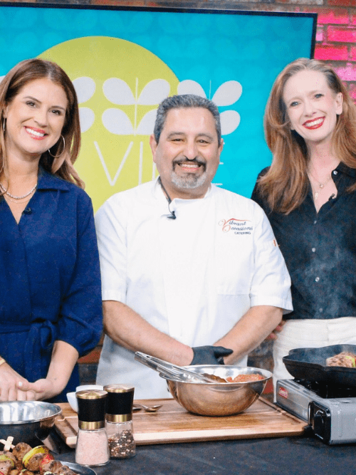 Grilled Steak Kabobs Recipe Three people, two women and one man in a chefs coat, stand smiling behind a kitchen counter with cooking ingredients and utensils, on a brightly lit TV set with a Viva logo in the background.